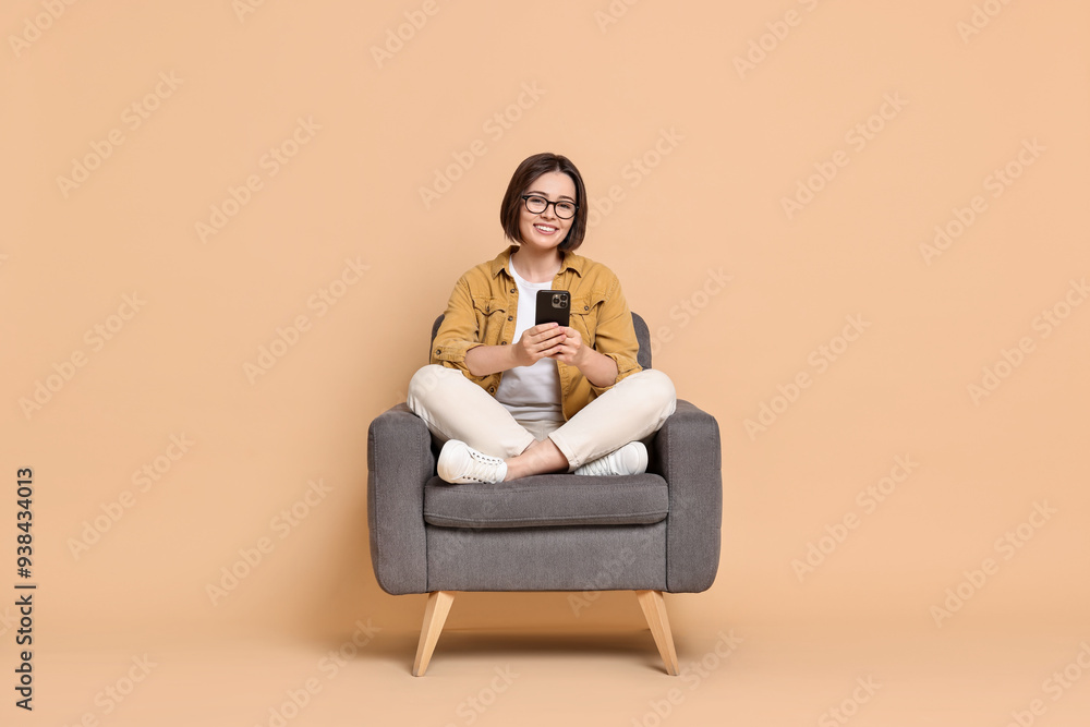 Smiling woman with smartphone sitting on armchair against beige background Smiling woman with smartphone sitting on armchair against beige background