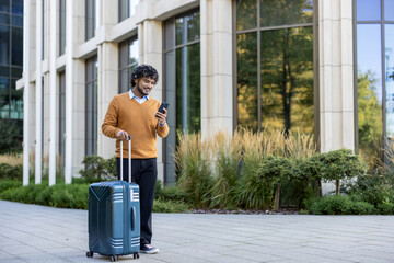 Smiling business traveler using smartphone while pulling suitcase in urban setting. Person wearing orange sweater walking outside office building. Captures concept of business travel