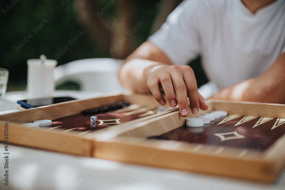 Close-up of a person enjoying a game of backgammon outdoors on a sunny day. The focus is on the hand and game pieces, creating a relaxed and strategic atmosphere.