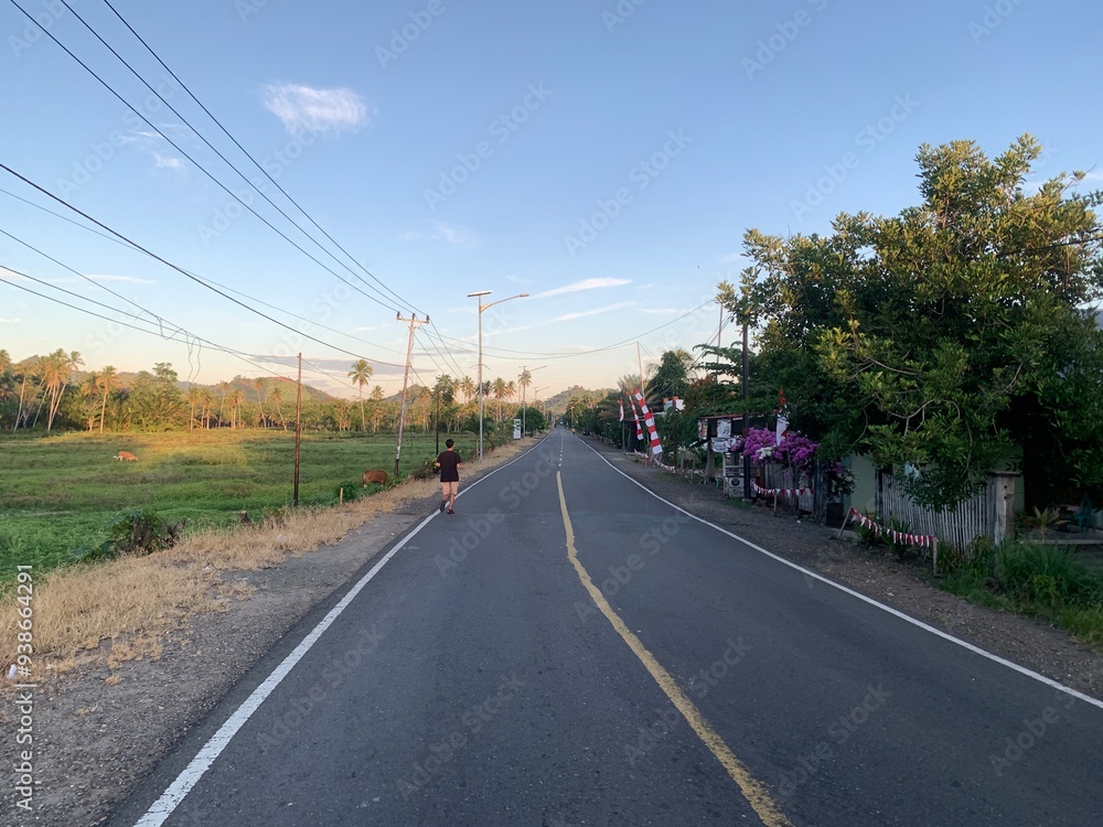 Man Running on a Rural Road at Dawn. A man runs on a straight rural road at sunrise in a tropical country. The road is surrounded by a green field, trees and houses. Man Running on a Rural Road at Dawn. A man runs on a straight rural road at sunrise in a tropical country. The road is surrounded by a green field, trees and houses.