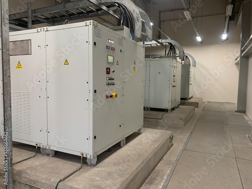 Industrial Equipment in a Factory. A row of industrial equipment, likely electrical transformers, are lined up in a factory setting. The equipment is white and has control panels with wires and cables
