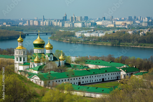 Peaceful monastery with golden domes and green roofs beside a serene river, juxtaposed against a backdrop of a modern cityscape with numerous buildings visible.