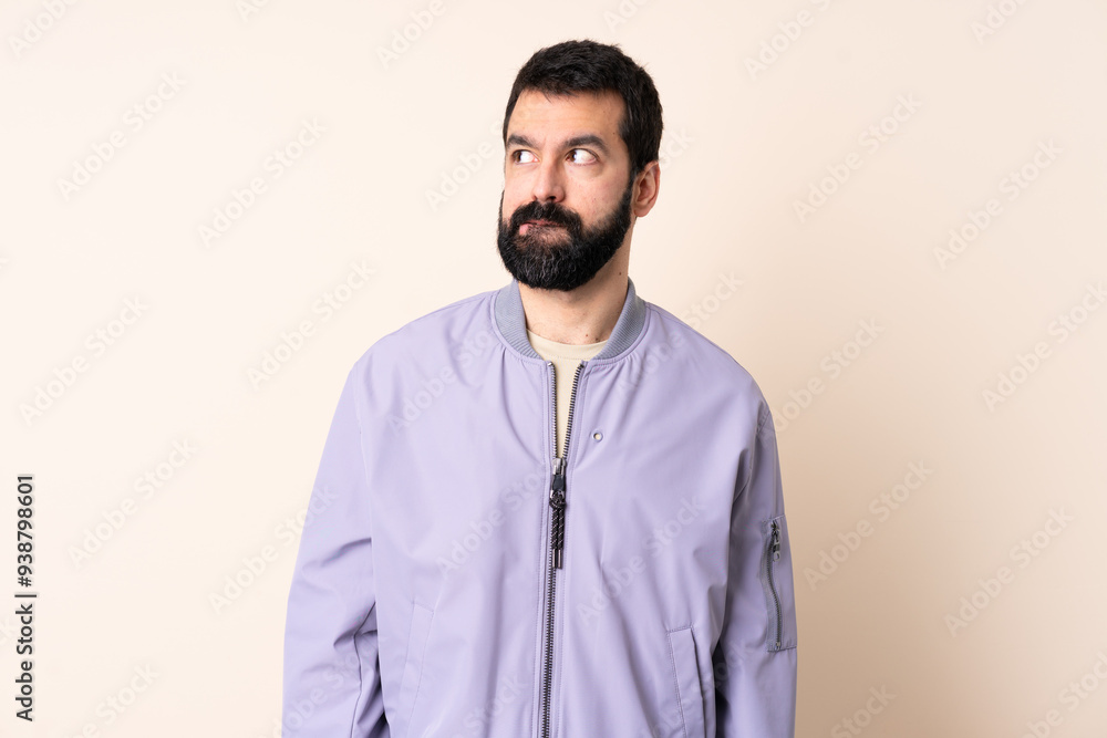 Caucasian man with beard wearing a jacket over isolated background having doubts while looking up Caucasian man with beard wearing a jacket over isolated background having doubts while looking up