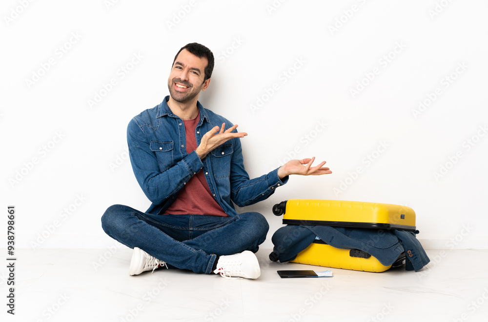Caucasian handsome man with a suitcase full of clothes sitting on the floor at indoors extending hands to the side for inviting to come Caucasian handsome man with a suitcase full of clothes sitting on the floor at indoors extending hands to the side for inviting to come