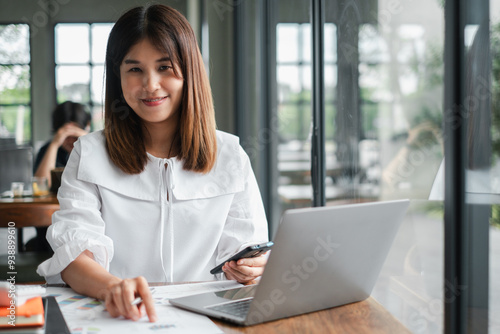 Smiling young woman working on a laptop in a modern office, holding a smartphone, and reviewing documents.