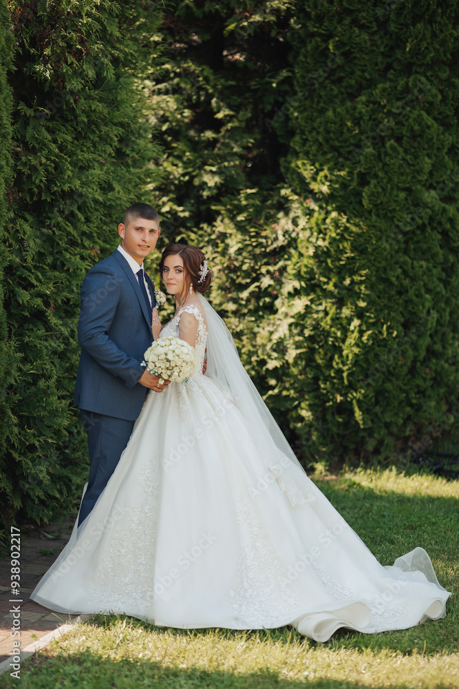 A bride and groom are posing for a picture in front of a bush. The bride is wearing a white dress and the groom is wearing a suit. The couple is holding a bouquet and a bouquet holder A bride and groom are posing for a picture in front of a bush. The bride is wearing a white dress and the groom is wearing a suit. The couple is holding a bouquet and a bouquet holder