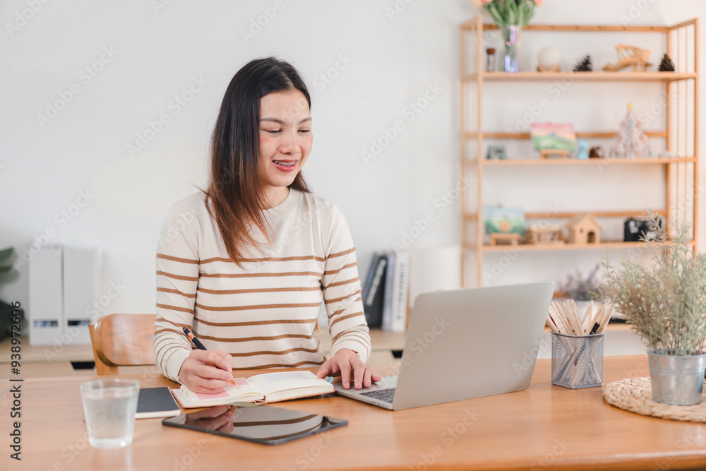 Smiling Asian woman typing on laptop while sitting at home, focused on work or study in a comfortable setting. Smiling Asian woman typing on laptop while sitting at home, focused on work or study in a comfortable setting.