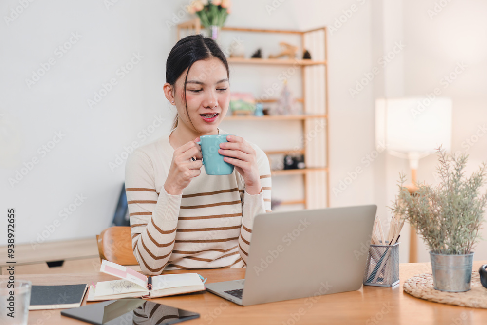 Asian woman sitting comfortably with a cup of hot beverage, smiling and enjoying a cozy moment in home office. Asian woman sitting comfortably with a cup of hot beverage, smiling and enjoying a cozy moment in home office.