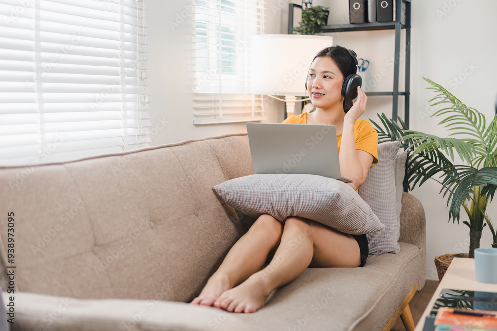 Smiling Asian woman relaxing on a cozy couch, wearing headphones, using a laptop, enjoying a comfortable home. Smiling Asian woman relaxing on a cozy couch, wearing headphones, using a laptop, enjoying a comfortable home.