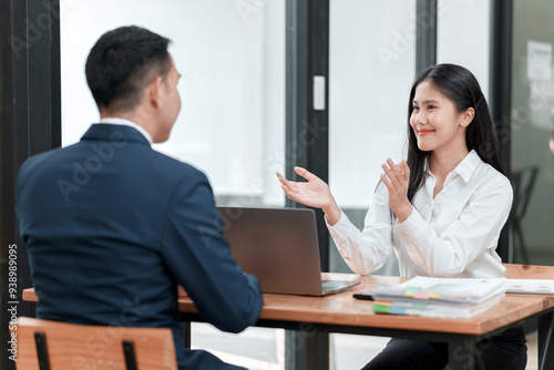 A man and a woman are sitting at a desk with a laptop in front of them. The woman is smiling and the man is looking at her. The laptop is open and there are papers on the table