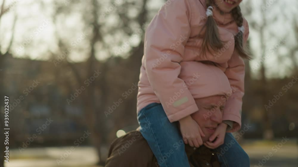A little girl in a pink cap and jacket sits joyfully on her dad's neck, half of his head snugly tucked in her jacket A little girl in a pink cap and jacket sits joyfully on her dad's neck, half of his head snugly tucked in her jacket