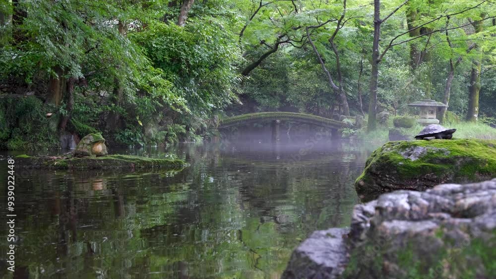A peaceful Japanese garden with a small, arched, moss-covered bridge spanning a pond with gentle ripples, surrounded by dense, lush greenery, trees, and plants. Zen and tranquil atmosphere. Gifu Park 
