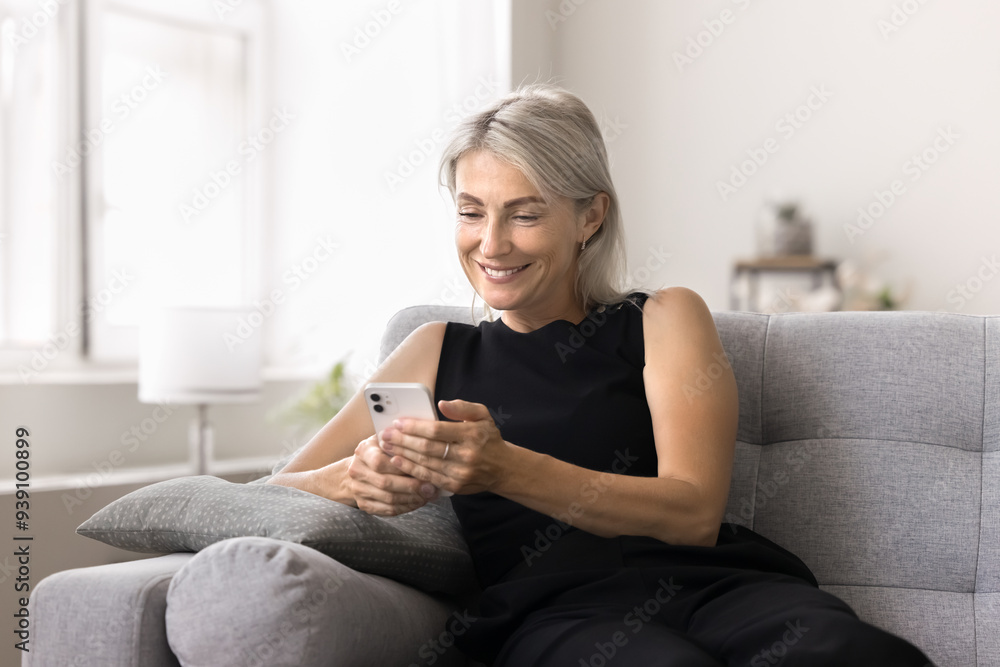 Mature woman using smartphone seated on couch at home, managing tasks, scheduling activities, enjoy on-line shopping, editing images, or creating content for social media using new mobile application Mature woman using smartphone seated on couch at home, managing tasks, scheduling activities, enjoy on-line shopping, editing images, or creating content for social media using new mobile application