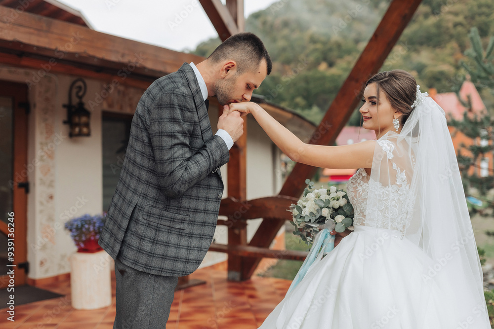 Fototapeta premium A man and a woman are kissing each other's hands. The man is wearing a suit and tie, and the woman is wearing a white dress. They are standing outside in front of a building