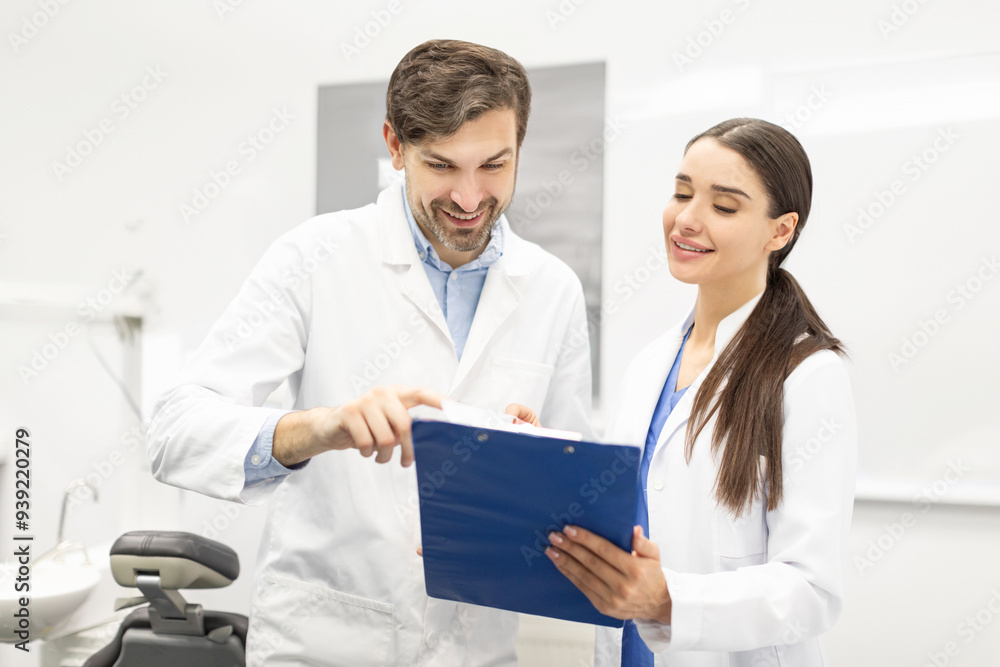 Male dentist doctor and his female assistant looking and discussing medical results on clipboard in modern dental clinic interior Male dentist doctor and his female assistant looking and discussing medical results on clipboard in modern dental clinic interior
