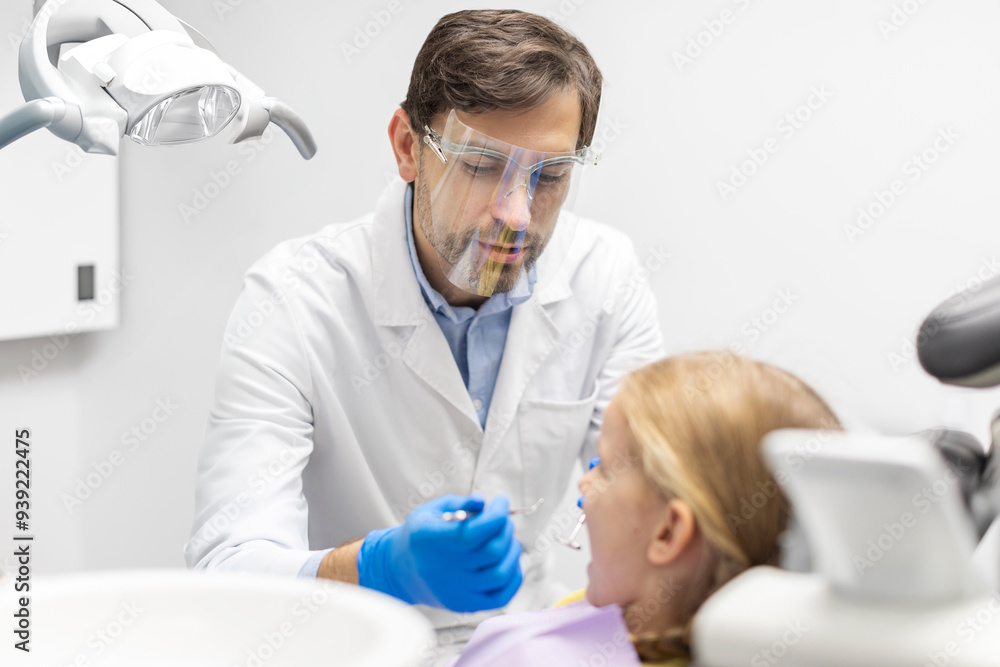 Obraz premium Little girl taking care of her teeth, sitting in chair while male dentist orthodontist in mask examining dental cavity