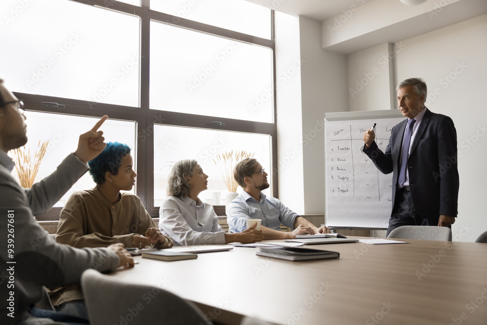 Group of professionals participating in meeting or workshop. Older presenter standing near flip chart give presentation interact with participants, attendee ask questions, share opinion or solutions Group of professionals participating in meeting or workshop. Older presenter standing near flip chart give presentation interact with participants, attendee ask questions, share opinion or solutions