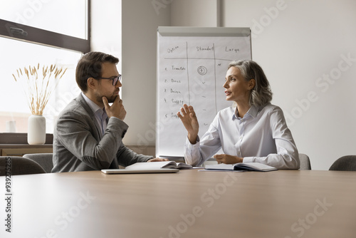 Two professionals met in conference room engaged in business-related discussion, mature female explaining or emphasizing point, while man listening attentively. Strategizing, problem solutions search