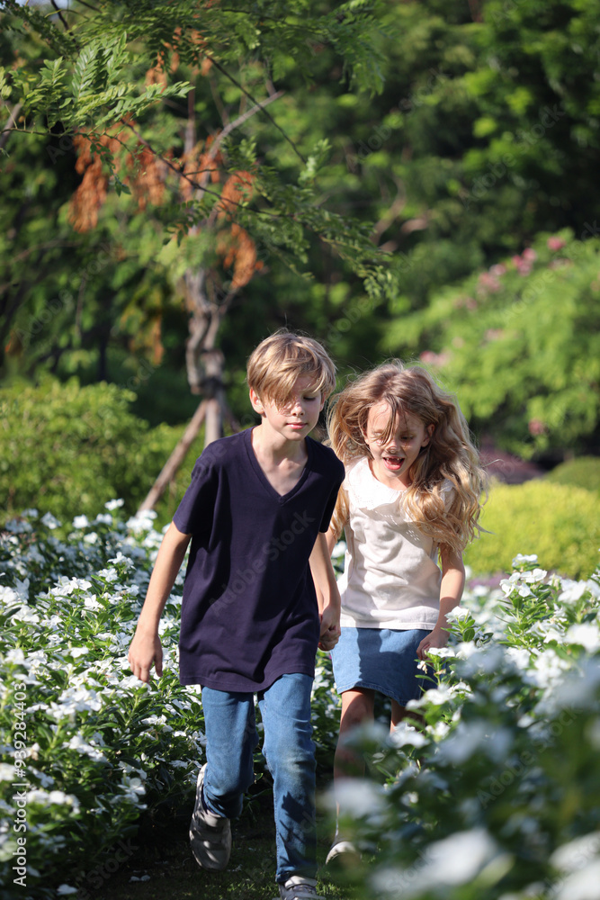 Fototapeta premium Happy family, brother and sister running through beautiful flower garden together. Joyful kids, boy and girl playing outdoor at public park. Children enjoy spend time outside. Sibling plays together.