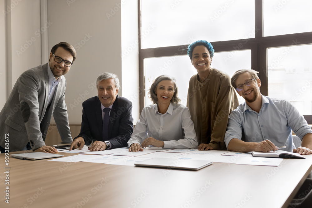 © fizkes - Five multiracial teammates gathered in conference room, smiling at camera, engaged in collaborative project, exuding professionalism, take part in productive and effective teamwork. Unity, business