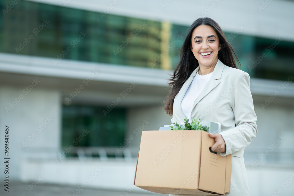 A woman dressed in a white suit is joyfully carrying a box filled with plants and personal belongings outside a contemporary building, showcasing a blend of business and nature. A woman dressed in a white suit is joyfully carrying a box filled with plants and personal belongings outside a contemporary building, showcasing a blend of business and nature.
