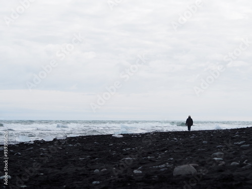 A solitary figure walks along the black sand beach, the dark sands and turbulent waters creating a mood of isolation and contemplation under the expansive gray sky - Iceland.