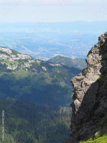 An expansive view over a lush green valley towards distant mountain ranges and a far-off city, showcasing the vastness and beauty of the landscape from a high vantage point.