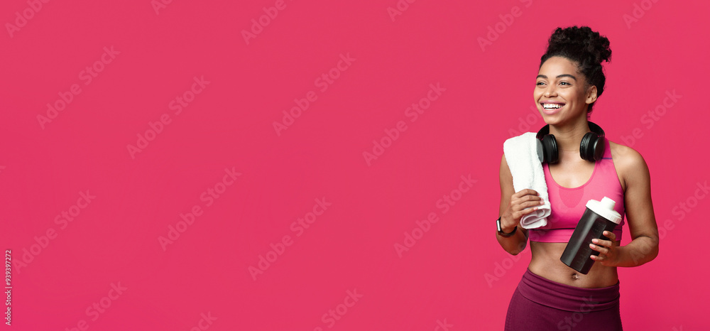 Cheerful african fitness girl with towel on her shoulder and wireless headset holding bottle with protein, looking at copy space Cheerful african fitness girl with towel on her shoulder and wireless headset holding bottle with protein, looking at copy space