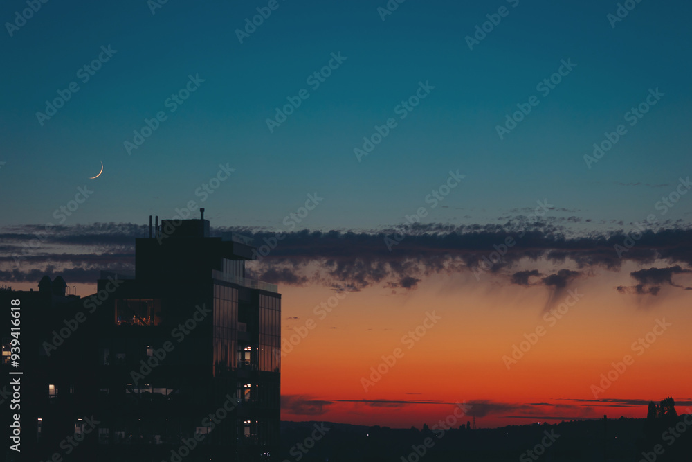 Fototapeta premium Cityscape with wonderful varicolored vivid dawn and the new moon. Amazing dramatic multicolored sky above dark silhouettes of city buildings at sunset. Atmospheric sunrise in overcast weather.