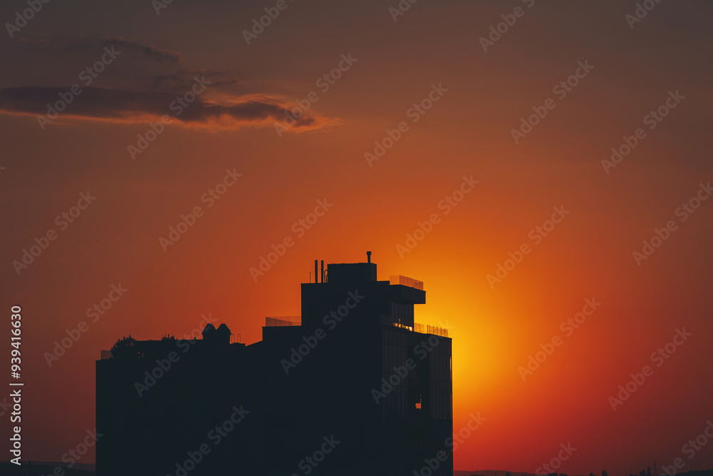 Fototapeta premium A stunning sunset paints the sky as the sun descends into clouds with a distant building in the foreground. The scene captures the beauty of urban and natural elements harmoniously blending at dusk.