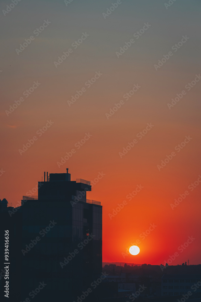 Obraz premium A stunning sunset paints the sky as the sun descends into clouds with a distant building in the foreground. The scene captures the beauty of urban and natural elements harmoniously blending at dusk.