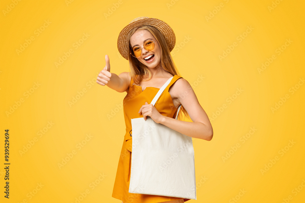 Stylish summer teen girl holding white canvas eco tote bag and showing thumb up, wearing straw hat and sunglasses, posing over yellow background Stylish summer teen girl holding white canvas eco tote bag and showing thumb up, wearing straw hat and sunglasses, posing over yellow background