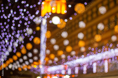 A vibrant blur of festive city lights, featuring soft white, blue, and yellow bokeh, illuminates the night. The colorful decorations create a magical, dreamlike atmosphere in an urban setting.