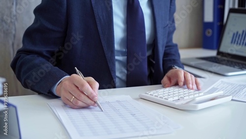 Businessman accountant with blue suit is using white calculator and taking notes with a silver pen. Taxes and audit concept