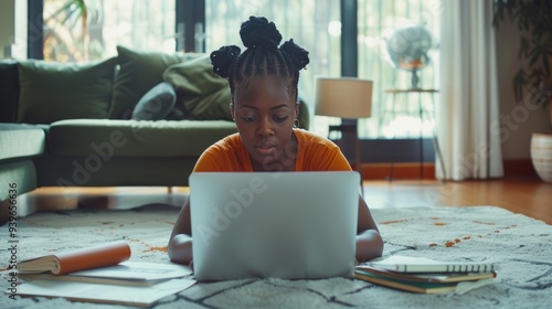 A young black woman is lying on the floor of her living room, working from home with an open laptop in front of her and some papers next to it. photo