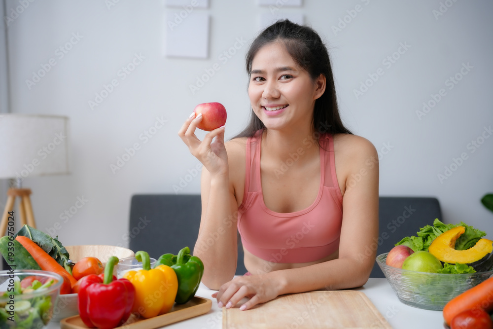 Young asian woman in sportswear is smiling while holding an apple in her kitchen with fresh vegetables on the table Young asian woman in sportswear is smiling while holding an apple in her kitchen with fresh vegetables on the table