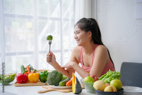 Young woman is smiling while holding a fork with a broccoli floret, surrounded by fresh vegetables on a table