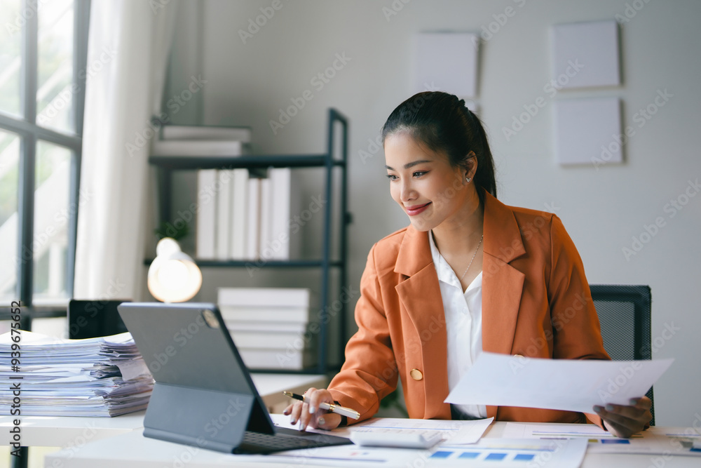 Asian businesswoman is smiling while working on a tablet and reviewing paperwork at her desk Asian businesswoman is smiling while working on a tablet and reviewing paperwork at her desk