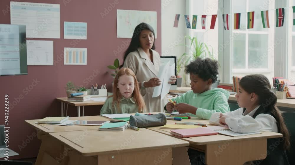 Medium shot of cheerful young female English teacher walking around classroom, handing out test sheets to group of diverse schoolchildren. Posters with grammar rules and country flags decorating walls Medium shot of cheerful young female English teacher walking around classroom, handing out test sheets to group of diverse schoolchildren. Posters with grammar rules and country flags decorating walls