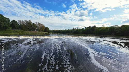 A rapid drone flyover capturing the flowing waters of Kuldiga Waterfall in Latvia, surrounded by lush greenery and a bright blue sky dotted with clouds. High quality 4k footage photo