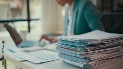 Accountant working diligently at a desk, surrounded by a large stack of invoices. Papers and documents clutter the workspace, reflecting a busy and demanding financial environment