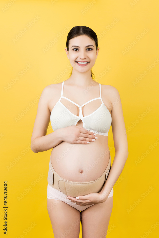 Portrait of pregnant woman in underwear wearing elastic bandage against pain in the back at yellow background with copy space. Orthopedic abdominal support belt concept Portrait of pregnant woman in underwear wearing elastic bandage against pain in the back at yellow background with copy space. Orthopedic abdominal support belt concept