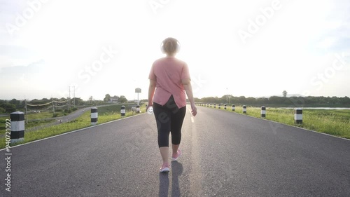A rear view of a middle-aged, overweight Asian woman walking for exercise on a road in the evening, wearing athletic wear and headphones, holding a water bottle, and embracing a healthy lifestyle