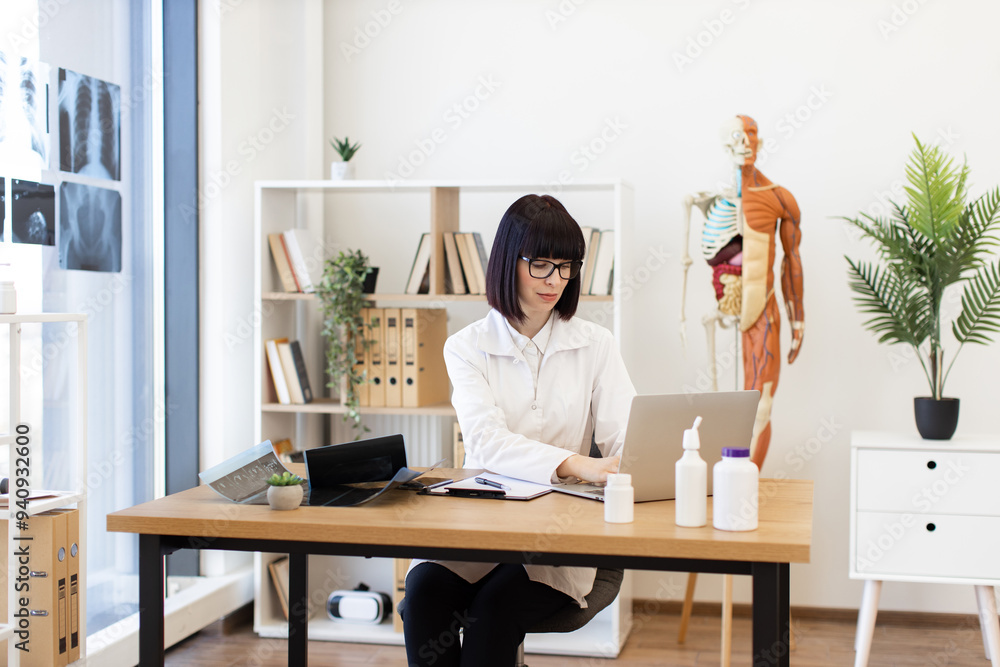 Female doctor focused on using laptop in modern medical office. Anatomical model, X-rays, and medical supplies on desk emphasize professional healthcare environment. Female doctor focused on using laptop in modern medical office. Anatomical model, X-rays, and medical supplies on desk emphasize professional healthcare environment.