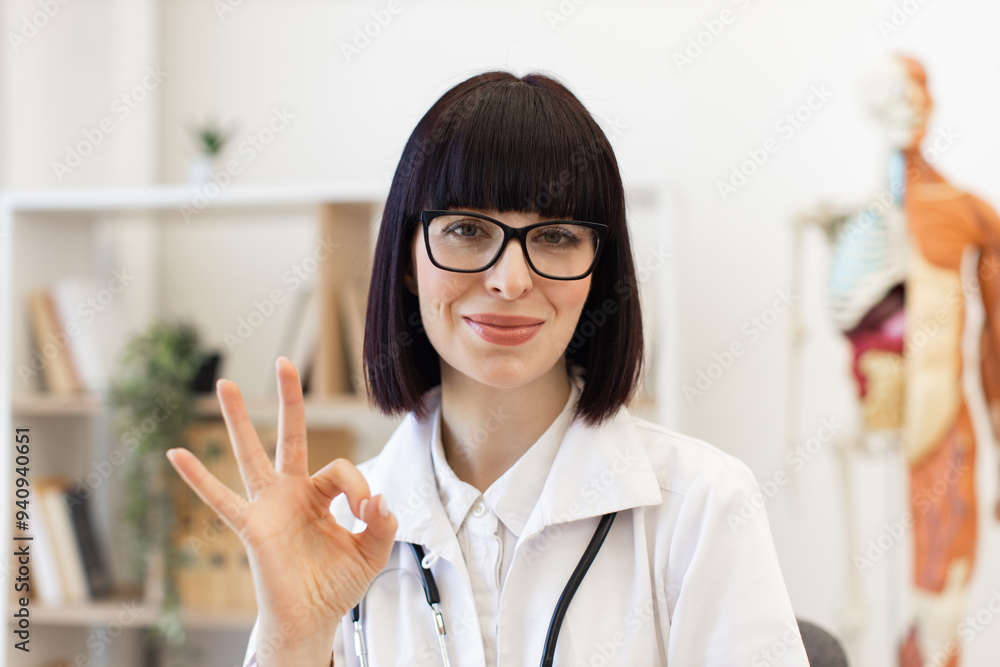 Smiling female doctor wearing glasses and white coat, giving ok sign with hand, expressing confidence and positivity in modern medical office. Smiling female doctor wearing glasses and white coat, giving ok sign with hand, expressing confidence and positivity in modern medical office.