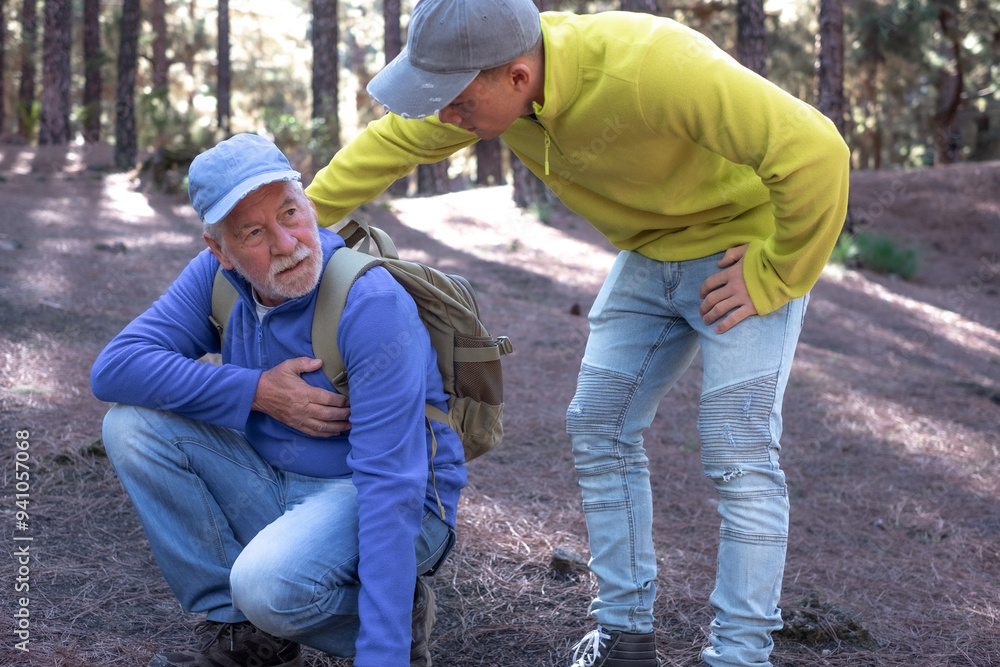 Old senior man hiking in the mountains with his young nephew pauses to catch his breath, touching his heart. Elderly grandfather feeling unwell and his grandson assists him Old senior man hiking in the mountains with his young nephew pauses to catch his breath, touching his heart. Elderly grandfather feeling unwell and his grandson assists him