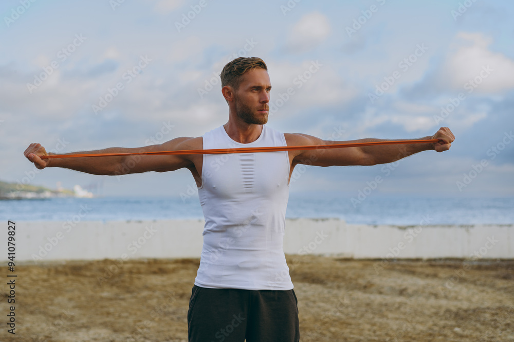 Young strong sporty athletic fit sportsman man he wear sports clothes using elastic rubber band for hands warm up training at sunrise sun dawn over sea beach outdoor on seaside in summer day morning. Young strong sporty athletic fit sportsman man he wear sports clothes using elastic rubber band for hands warm up training at sunrise sun dawn over sea beach outdoor on seaside in summer day morning.