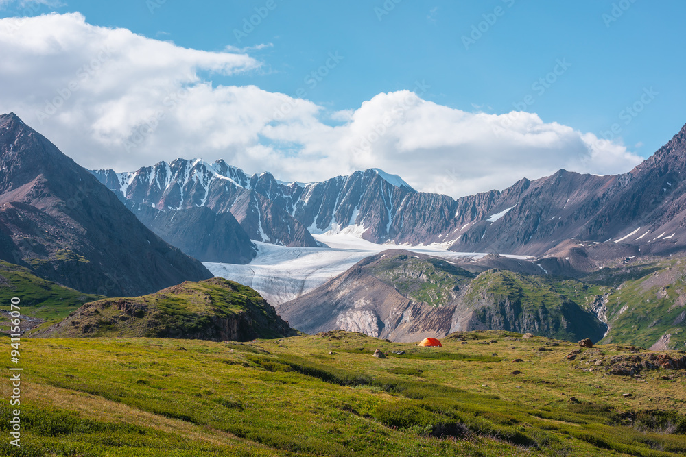 Obraz premium Colorful landscape with orange tent in alpine valley with view to big glacier tongue and large snow-capped mountain range under clouds in blue sky. Green hills and rocks against ice and sheer crags.