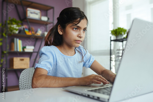 Young girl concentrating on laptop while sitting at desk in modern room with shelves and plants in background displaying engaged expression on face