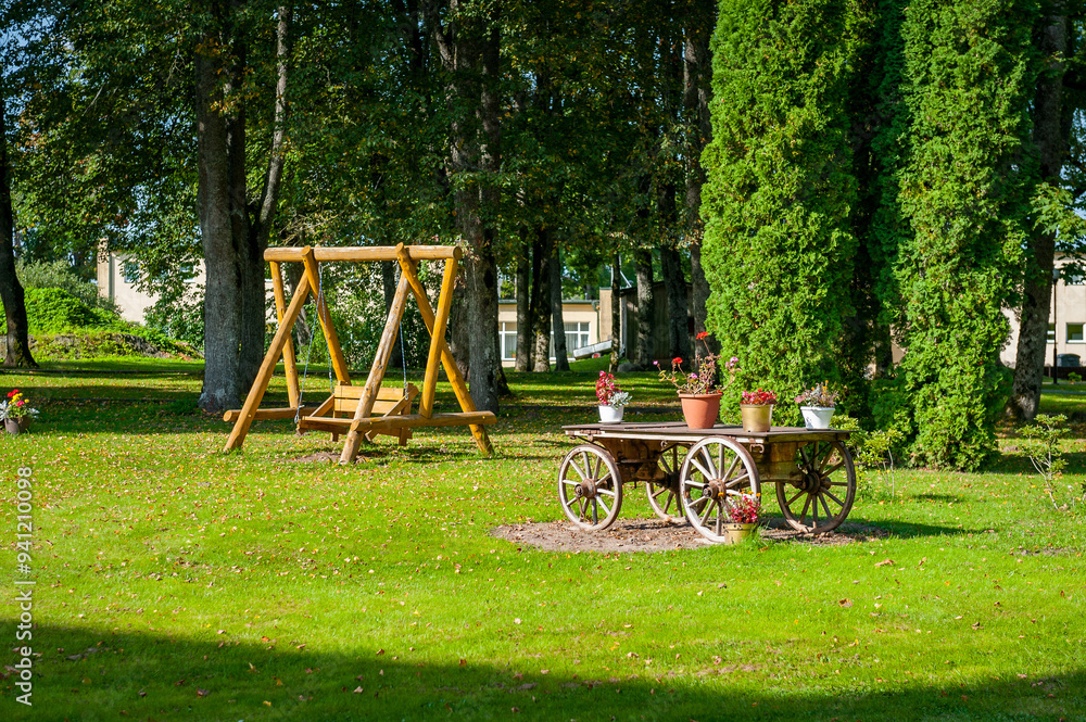 Wooden cart with flowers against the background of garden with wooden swing bench. Wooden cart with flowers against the background of garden with wooden swing bench.